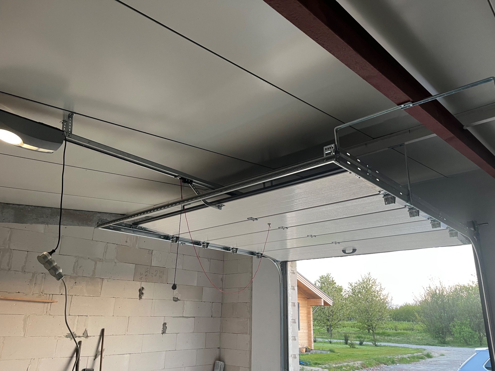 Widok od wewnątrz garażu na częściowo zamontowaną segmentową bramę garażową z widocznym mechanizmem podnoszenia i niezabudowaną ścianą z bloczków betonowych w tle.