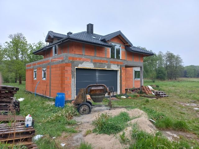 Budowa domu w trakcie realizacji: widoczne ściany z czerwonej cegły, zamontowane ramy okienne, szara brama garażowa, mieszalnik do betonu i materiały budowlane na zewnątrz.