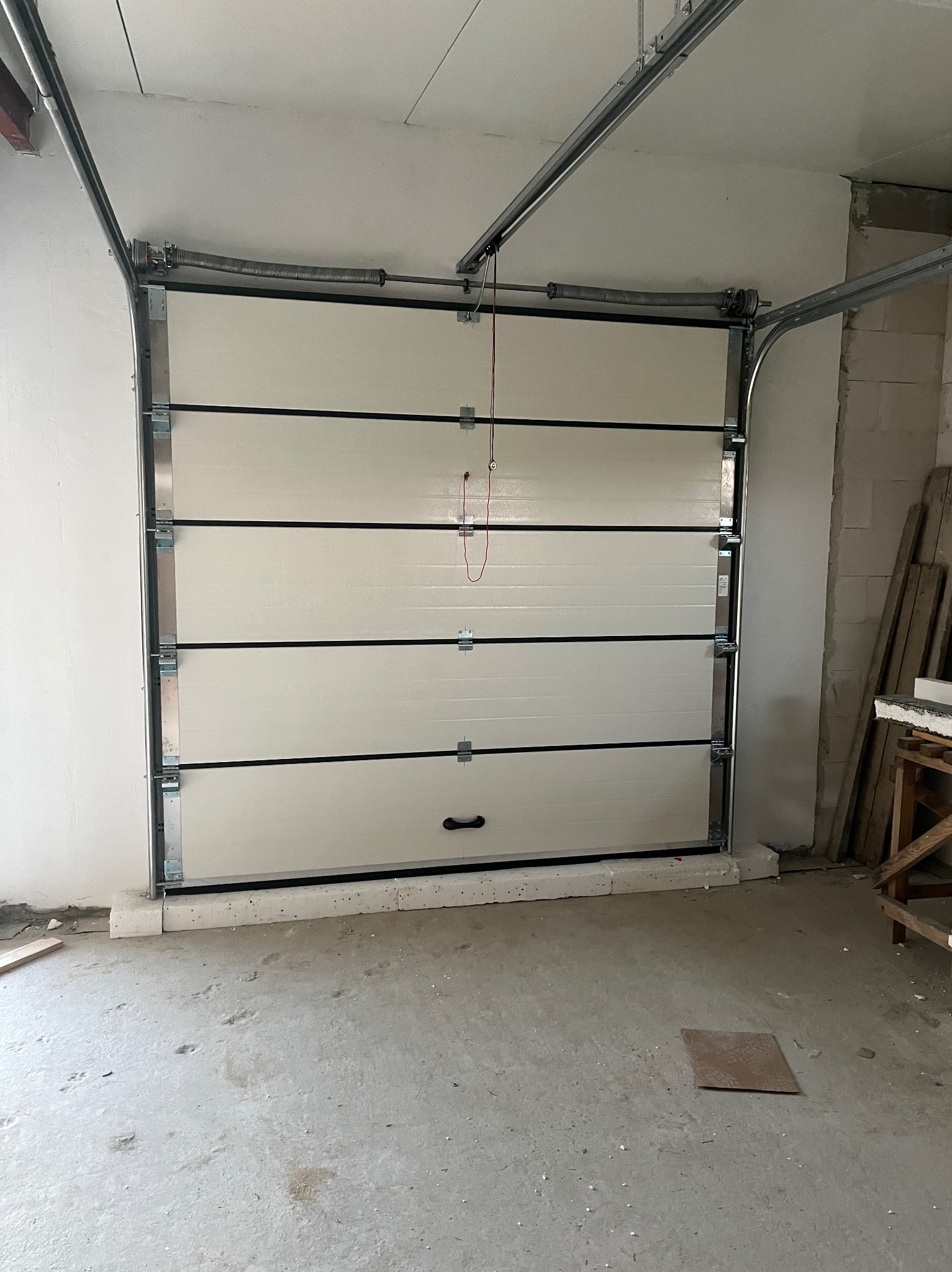 Zamknięta segmentowa brama garażowa w kolorze kremowym z czarnymi akcentami, widoczne sprężyny i prowadnice, surowy betonowy garaż w trakcie wykańczania.