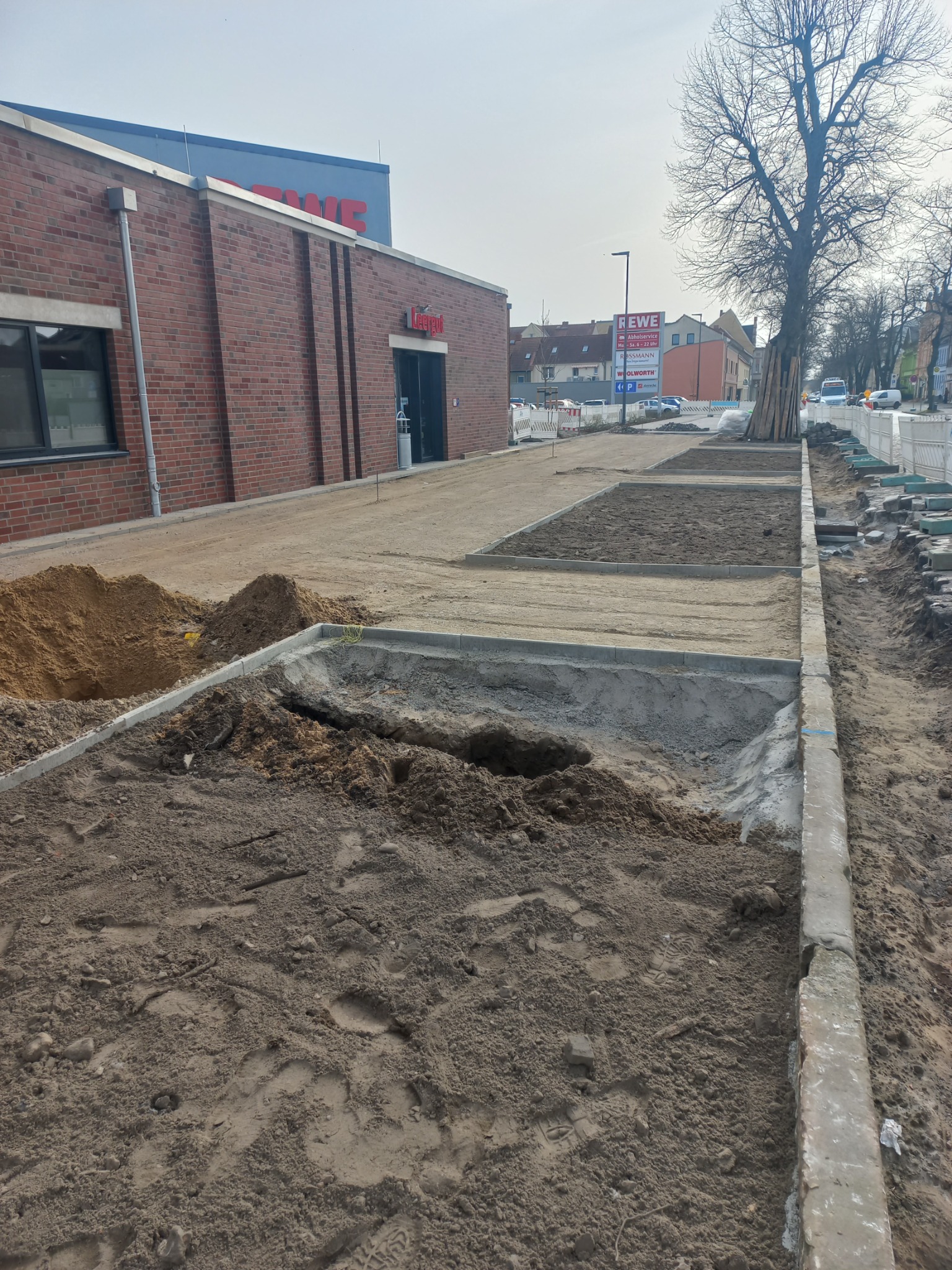 Przygotowanie podłoża pod chodnik z obrzeżami betonowymi przed budynkiem sklepu, widoczne pryzmy piasku i ziemi, fragmenty tymczasowego ogrodzenia.