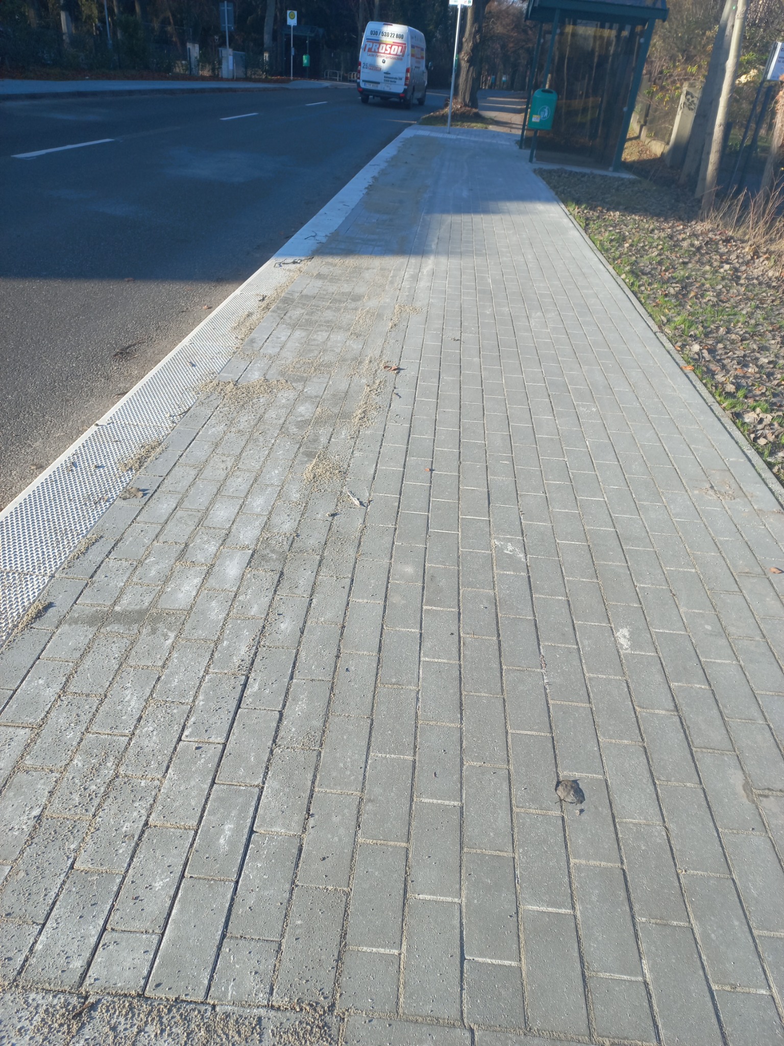 Świeżo ułożony chodnik z szarej kostki brukowej, częściowo jeszcze zanieczyszczony piaskiem, przylegający do ulicy z kratką ściekową i przystanku autobusowego w tle.