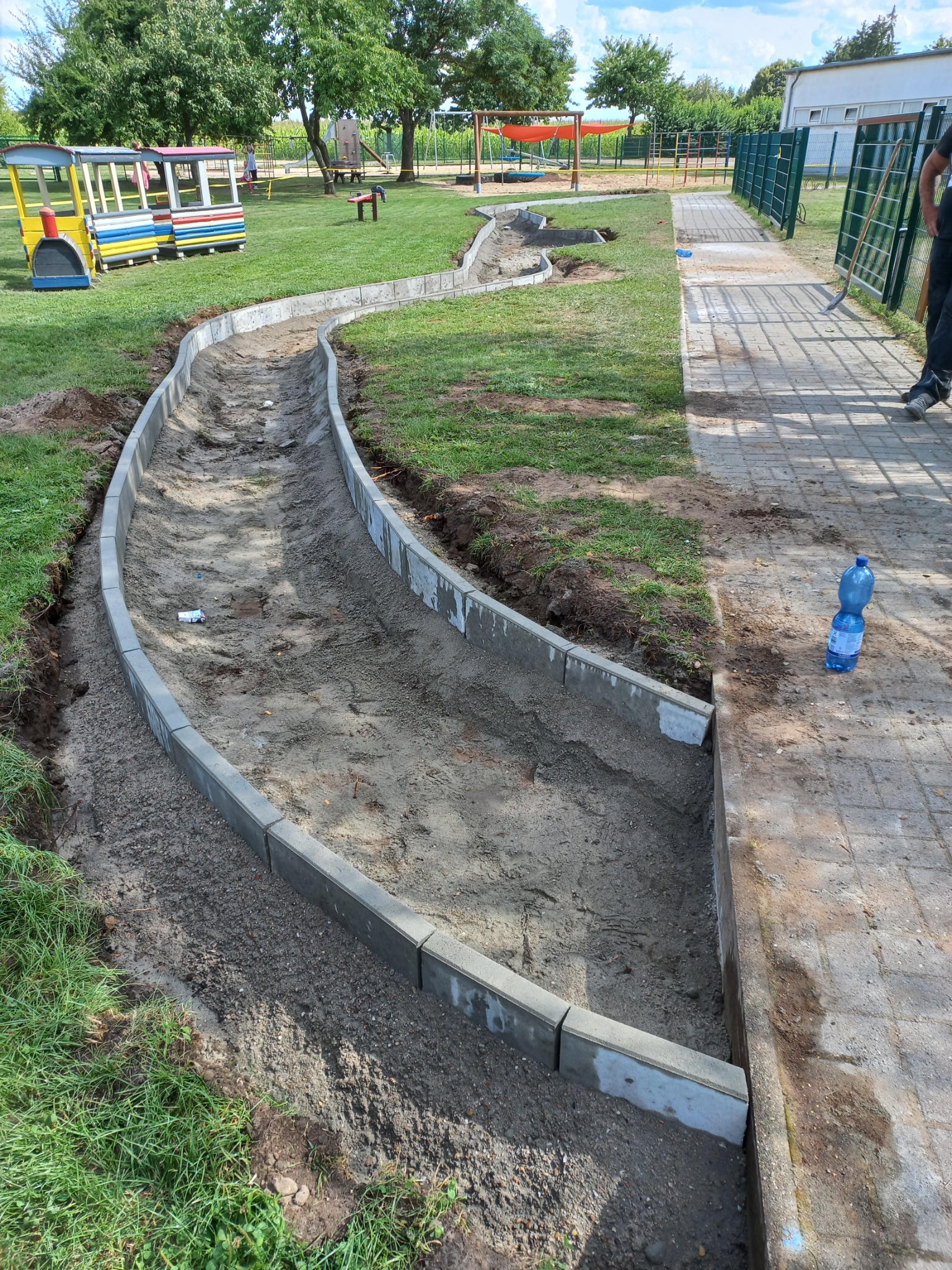Układanie obrzeży z kostki betonowej w kształcie zakrzywionej ścieżki na placu zabaw, widoczny teren przygotowany pod piasek, w tle kolorowy pociąg i elementy placu zabaw.