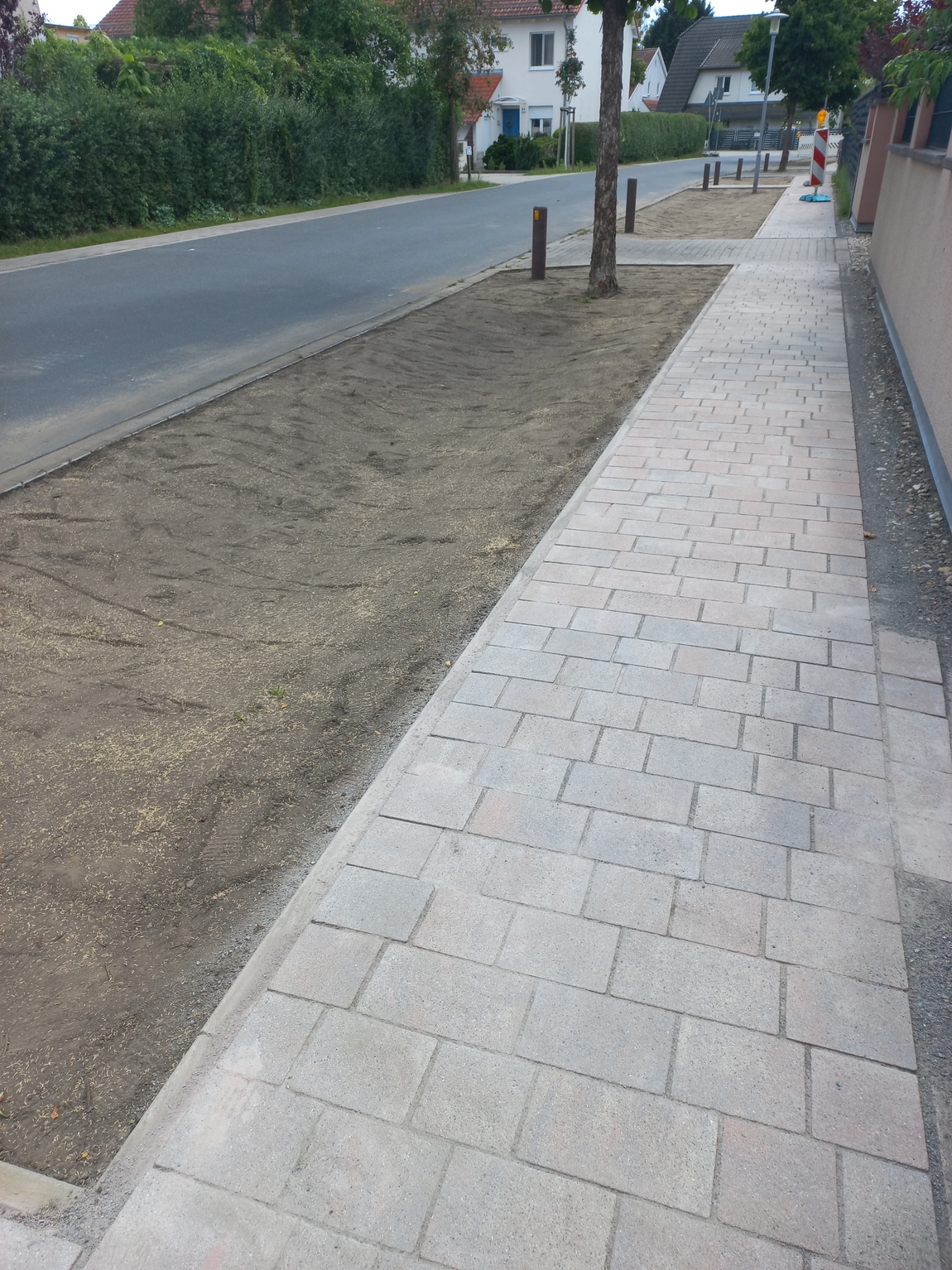 Nowo ułożona, jasnoszara kostka brukowa chodnikowa, przylegająca do drogi i przygotowanego podłoża pod trawnik z młodą trawą, oddzielona obrzeżem.