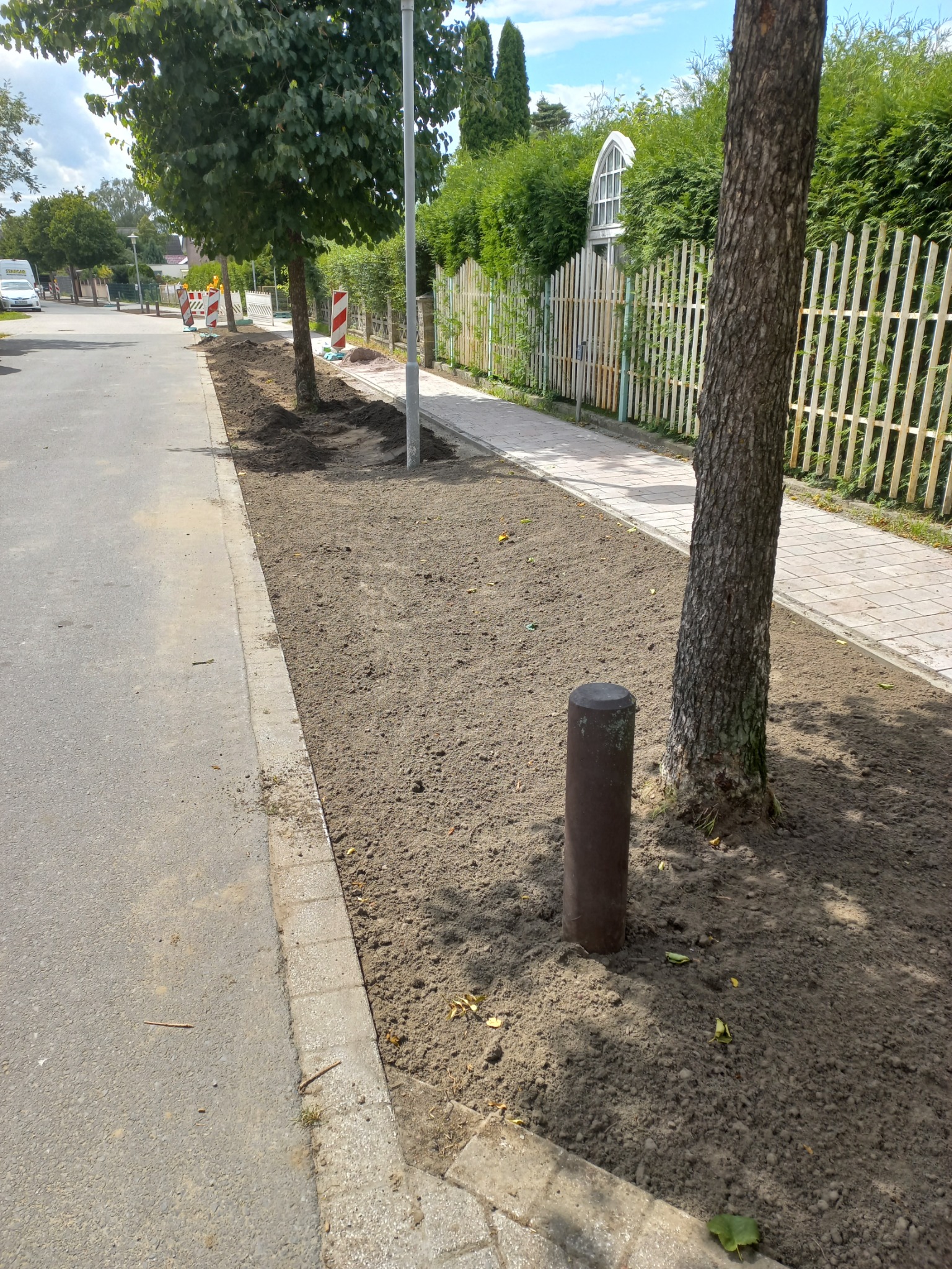 Przygotowany teren pod chodnik z obrzeżami, widoczne drzewa i słupki, w tle białe ogrodzenie i zabudowa.