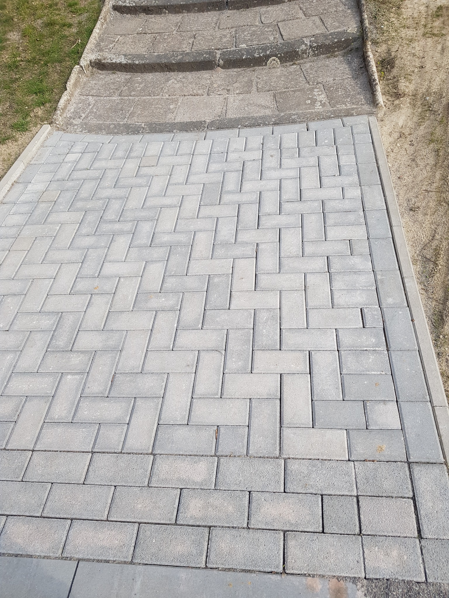 Ułożona z szarej kostki brukowej ścieżka w jodełkę, prowadząca do betonowych schodów na trawnik.