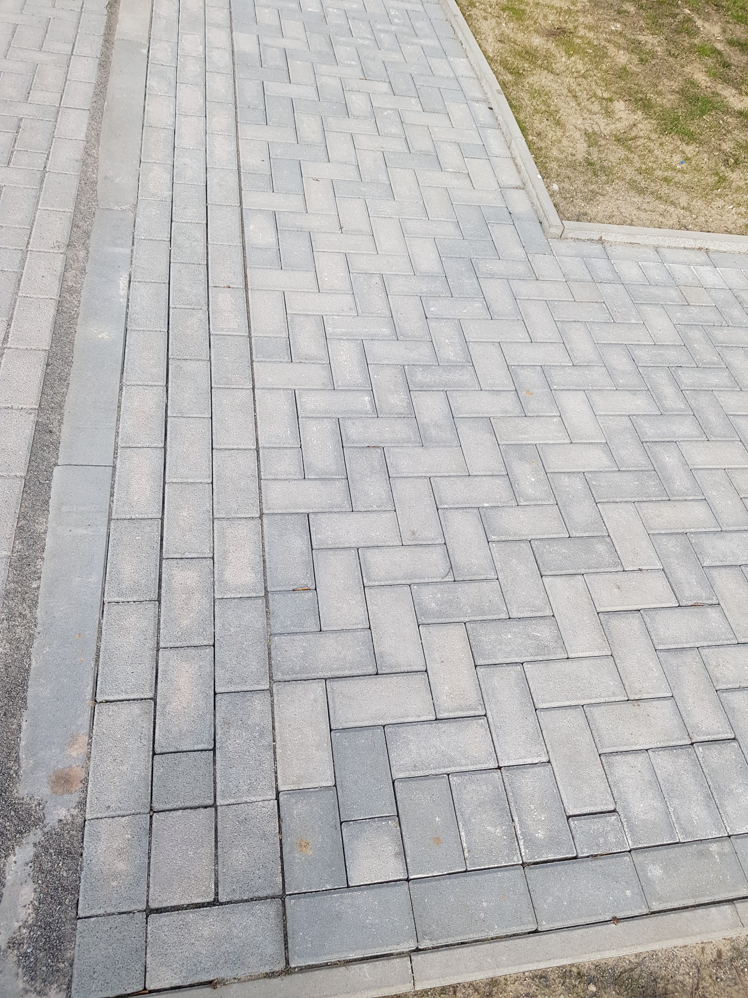Ułożona szara kostka brukowa w jodełkę, widok z góry na gotową nawierzchnię z obrzeżami, sąsiadującą z trawnikiem.