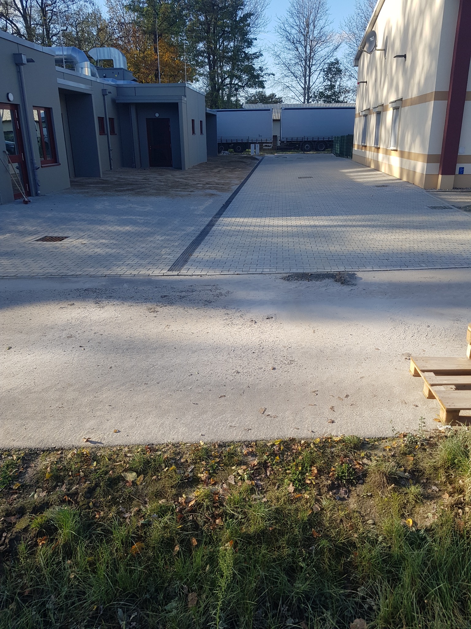 Szeroki widok na ułożoną szarą kostkę brukową na podjeździe między budynkami, z widocznymi studzienkami kanalizacyjnymi i zielenią w tle.