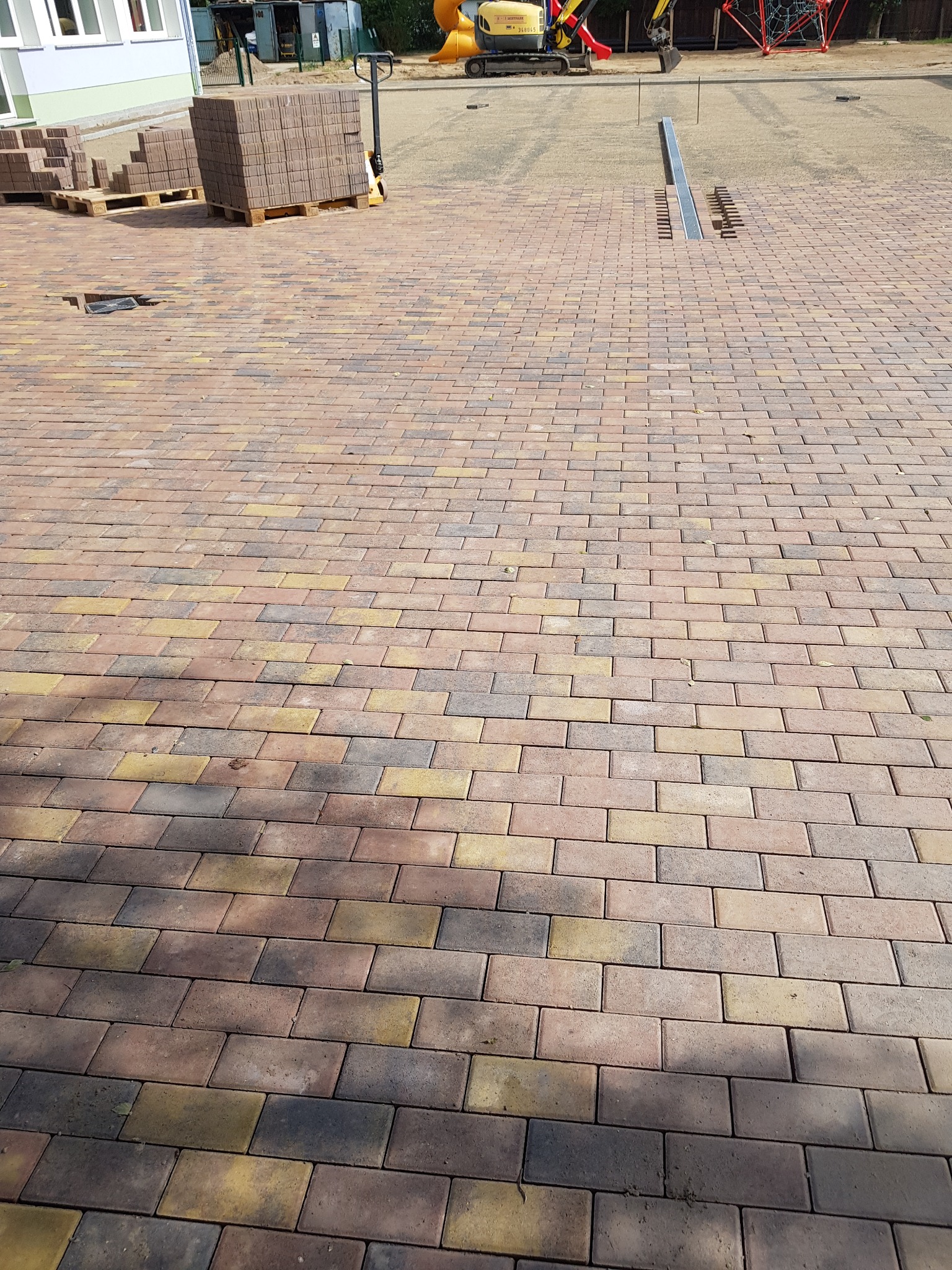 Ułożona kostka brukowa w odcieniach brązu, żółci i szarości, z paletami kostki w tle i widoczną linią odpływową oraz placem budowy.