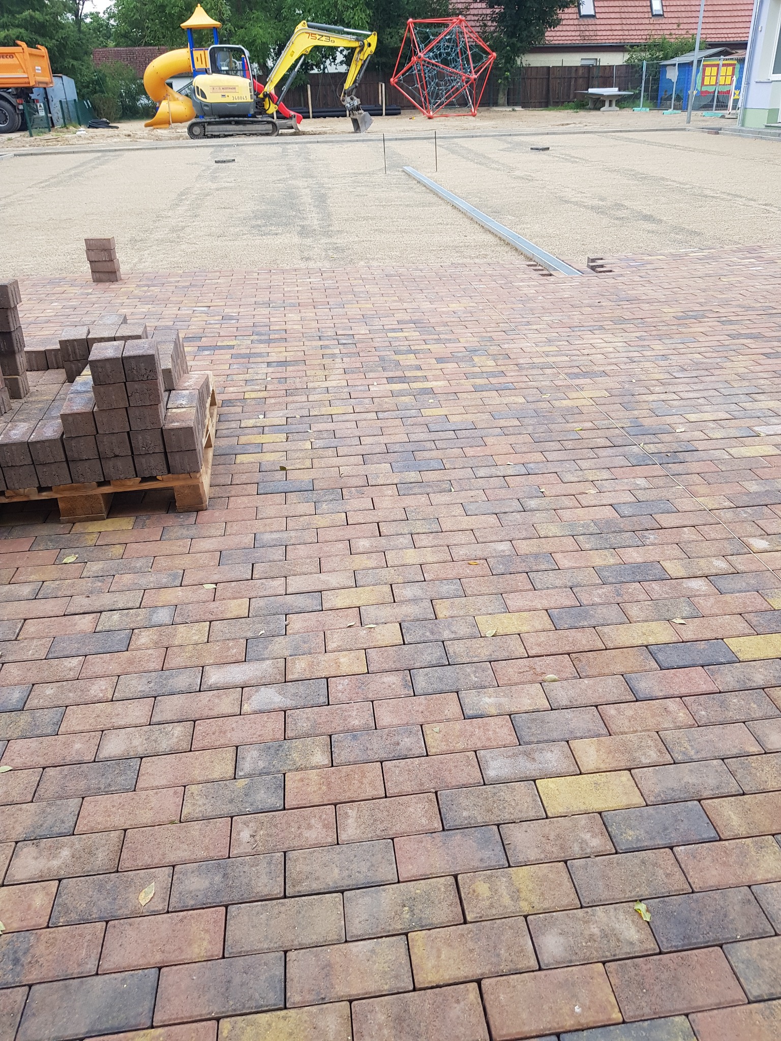 Układanie kostki brukowej na placu zabaw; widoczne palety z kostką, częściowo ułożona nawierzchnia z kostki w odcieniach brązu i żółci, w tle piasek, plac zabaw z żółtą zjeżdżalnią i koparką.