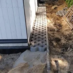 Montaż ażurowych płyt betonowych przy ścianie budynku, widoczny piasek i przygotowany teren pod dalsze prace.