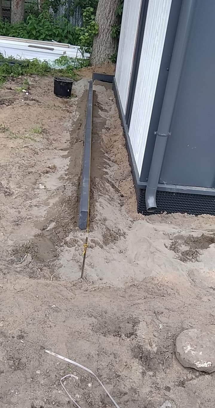 Przygotowanie podłoża pod obrzeża kostki brukowej wzdłuż ściany budynku z zamontowanym systemem rynnowym, widoczny wykop, wbity palik z naciągniętym sznurkiem wyznaczającym linię.