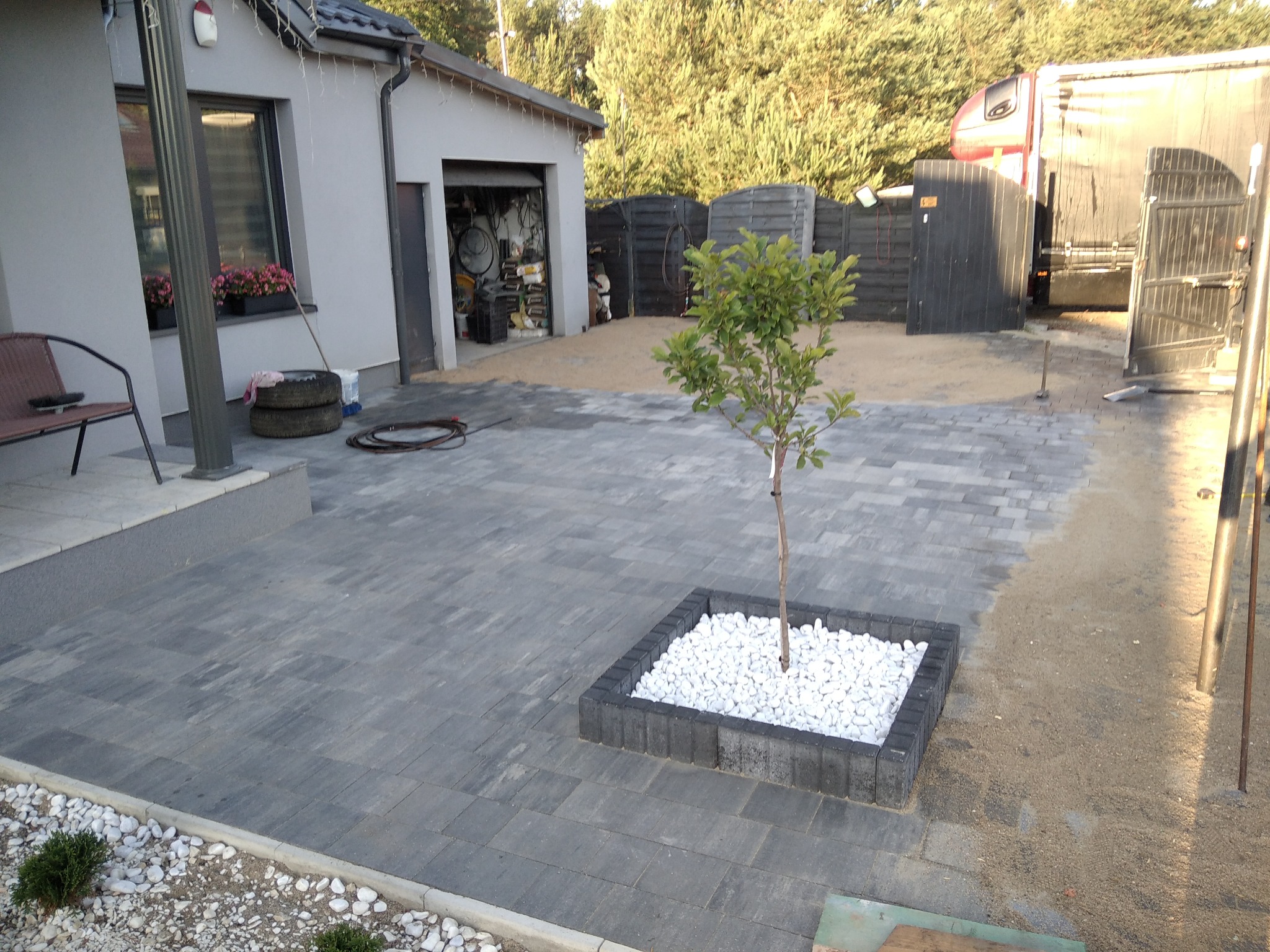 Ułożona szara kostka brukowa na podwórku z małym drzewkiem w kwadratowej obudowie wypełnionej białymi kamieniami, widoczny fragment domu z oknem, garaż z otwartymi drzwiami i zaparkowana ciężarówka.