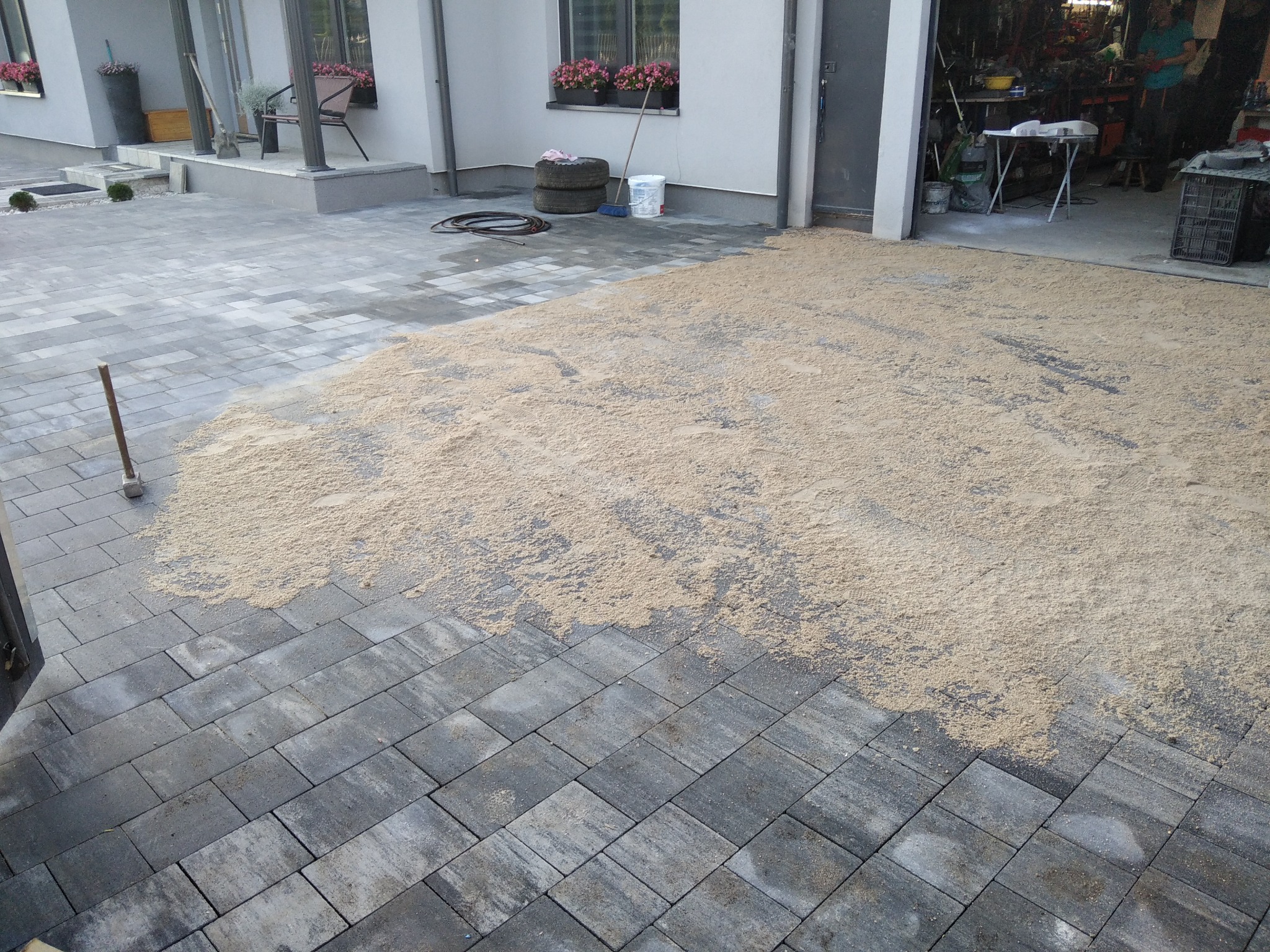 Układanie szarej kostki brukowej na podwórzu, częściowo zasypanej piaskiem, widok na dom z kwiatami w oknach i otwarty garaż.