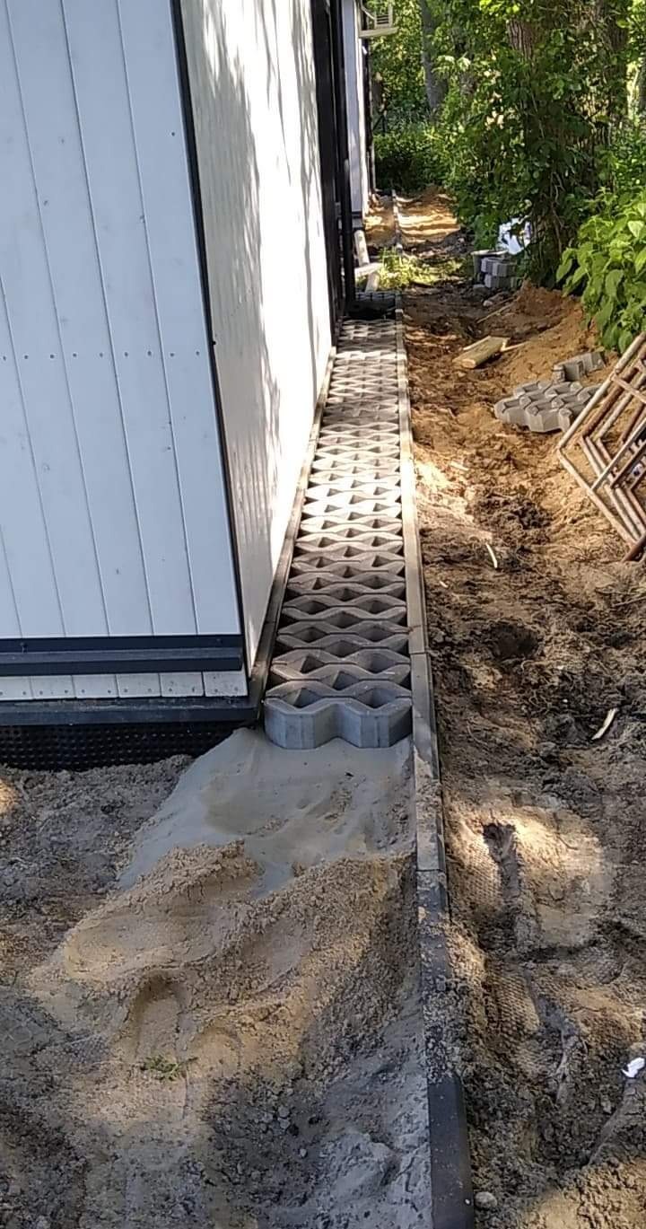 Montaż ażurowych płyt betonowych przy ścianie budynku, widoczny piasek i przygotowany teren pod dalsze prace.