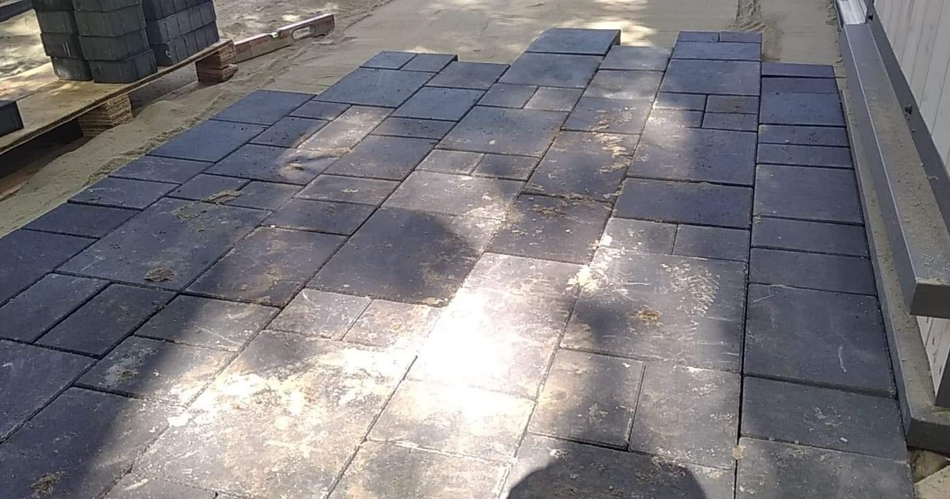 Ciemna kostka brukowa ułożona na piasku, widoczne nierówności i ślady piasku na powierzchni, paleta z kostką w tle, częściowo ułożona nawierzchnia.