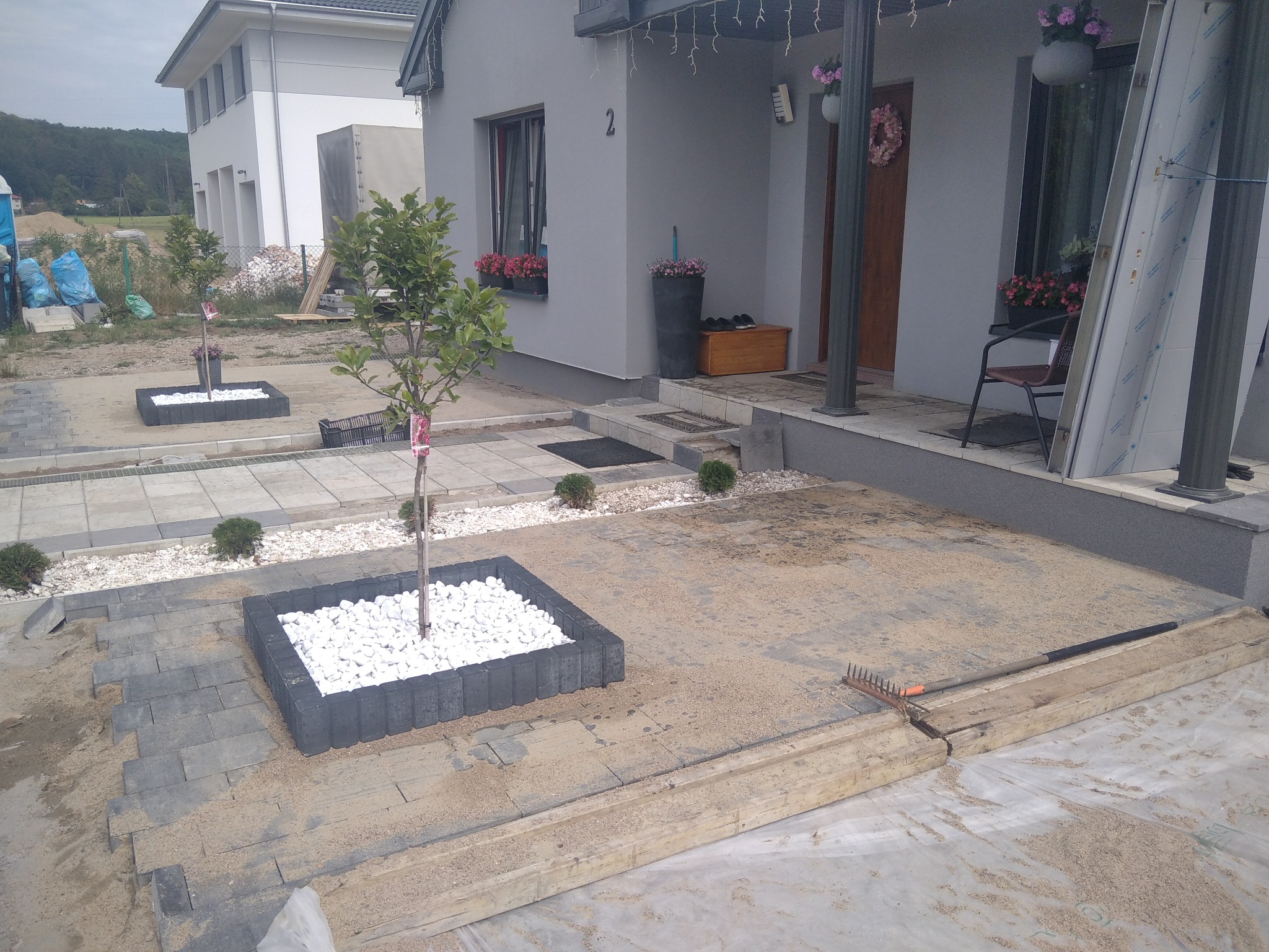 Ułożona kostka brukowa na posesji przed domem jednorodzinnym, widoczne rabaty z białym kamieniem i młodymi drzewkami, w tle dom z numerem 2 i elementy ogrodu.