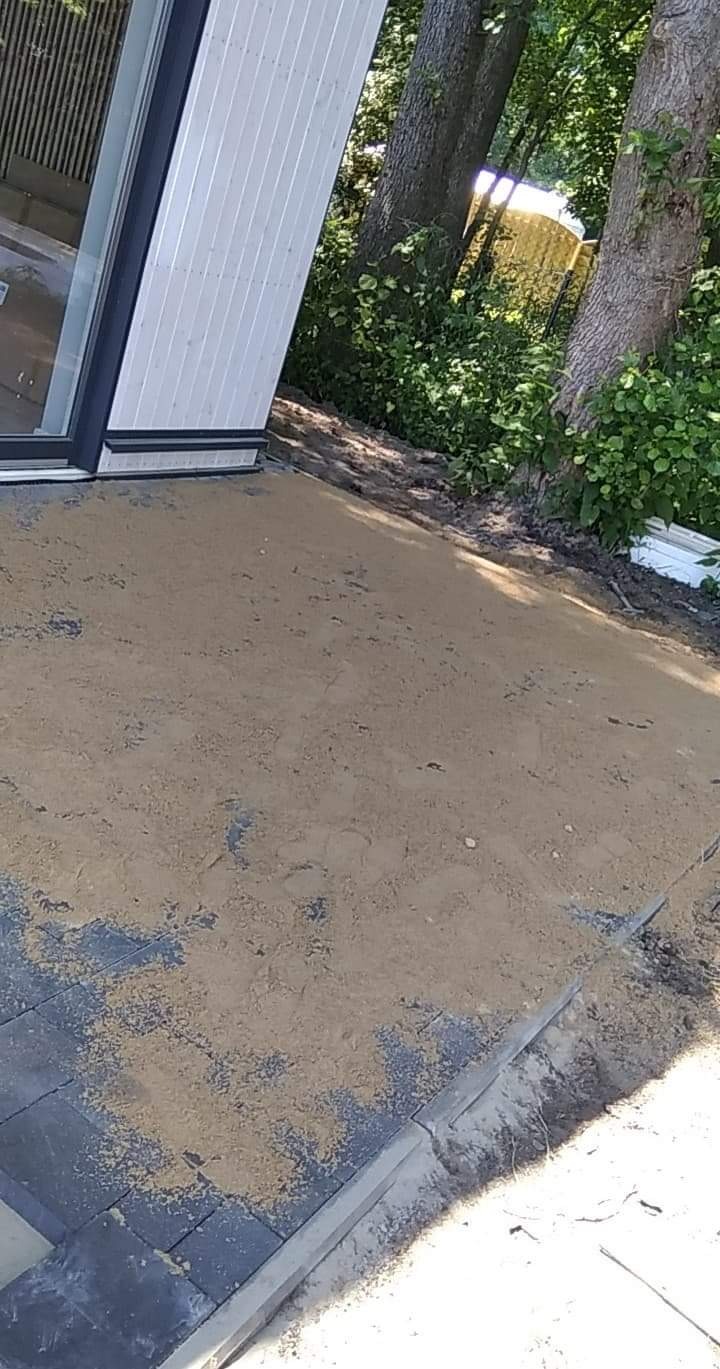 Przygotowanie podłoża pod kostkę brukową, podsypka piaskowa wyrównana obok budynku z szarą elewacją, widoczne fragmenty ułożonej kostki i krawężników.