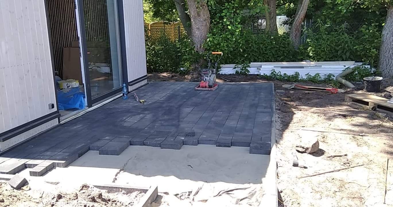 Układanie ciemnoszarej kostki brukowej na piasku przy nowym domu z przesuwnymi drzwiami, widoczny zagęszczarka wibracyjna i narzędzia.