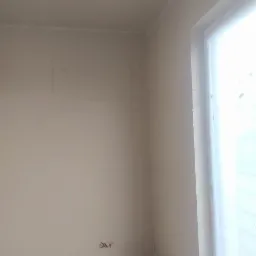 Wnętrze w trakcie prac wykończeniowych: ściany przygotowane pod malowanie, widoczne ślady po szpachlowaniu i gładziach, surowy beton na podłodze, wystające kable elektryczne, okno zasłonięte folią.