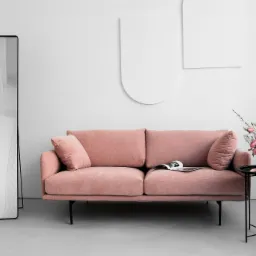 Jasnoróżowa kanapa z poduszkami, czarny stolik z bukietem różowych róż, wysokie lustro i minimalistyczne dekoracje ścienne w jasnym, minimalistycznym wnętrzu.