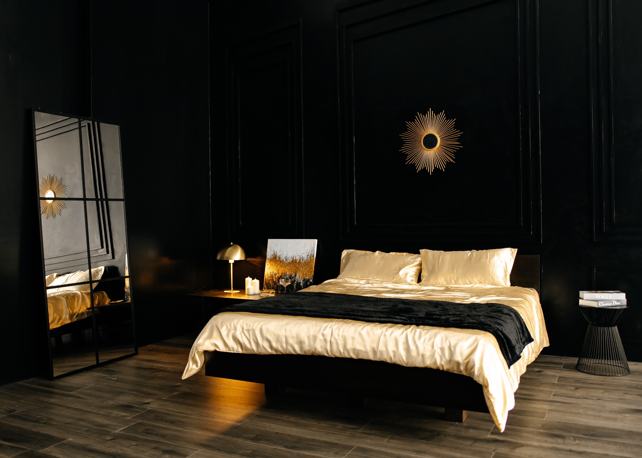 Elegancka sypialnia w czerni i złocie: łóżko z satynową pościelą, czarny pled, lustro w metalowej ramie, złota lampka nocna i dekoracja ścienna w kształcie słońca.