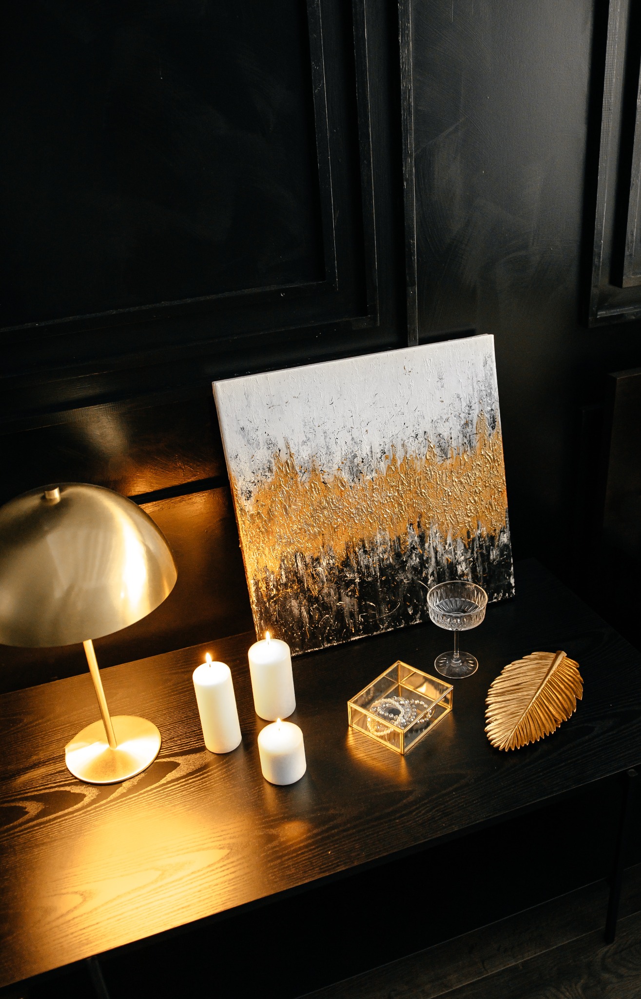 Martwa natura: abstrakcyjny obraz z fakturą złota, zapalone świece, lampka nocna, szklana szkatułka z biżuterią i dekoracyjny liść na czarnym stoliku.