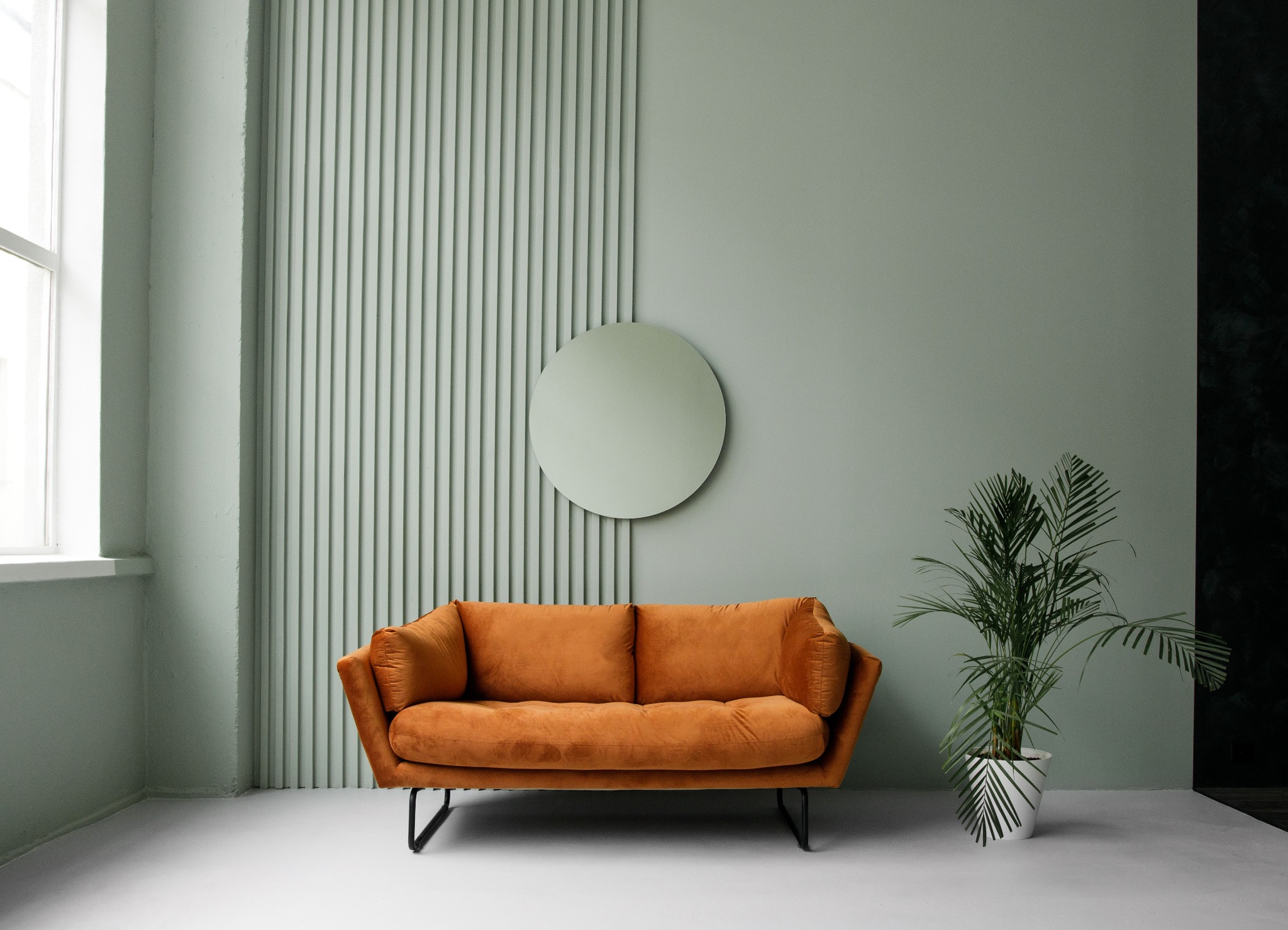 Pomarańczowa sofa na czarnych nóżkach, okrągłe lustro na ścianie w odcieniach zieleni, roślina doniczkowa w białej doniczce z czarnymi wzorami.