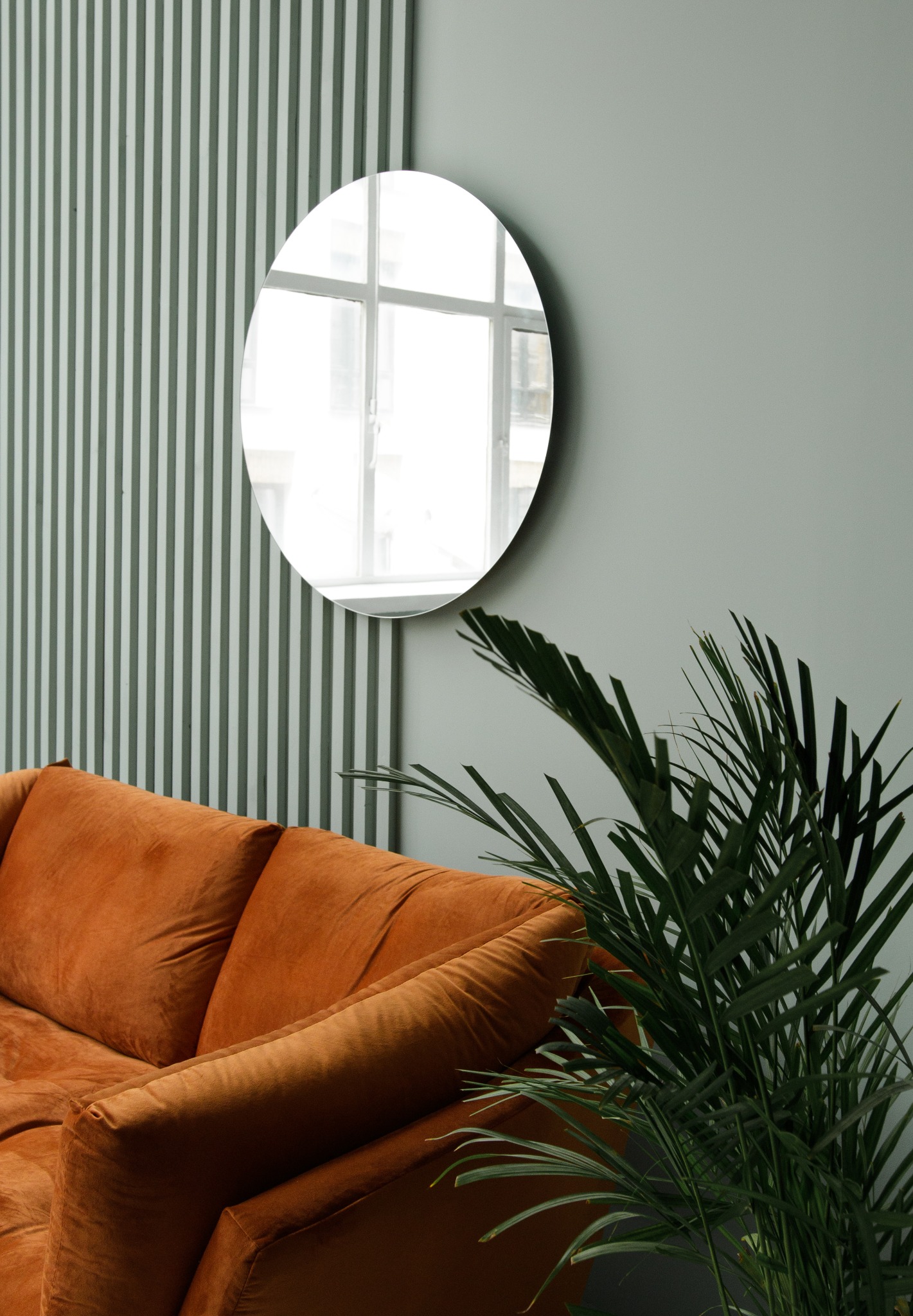 Wnętrze pokoju z pomarańczową sofą, okrągłym lustrem i zieloną rośliną na tle ściany w pionowe pasy.