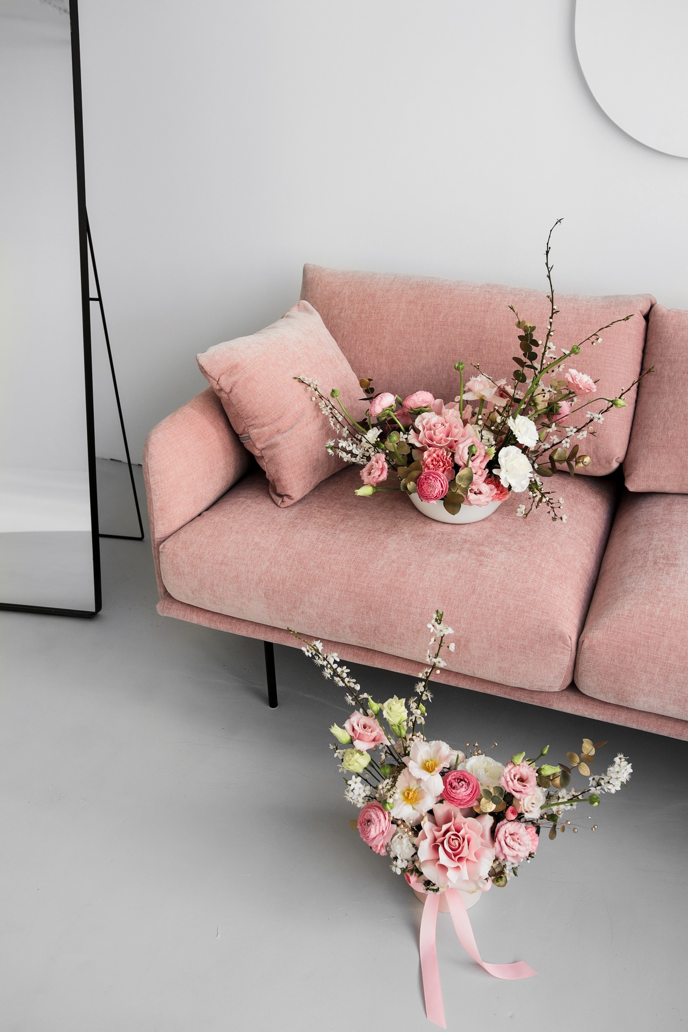 Kompozycja kwiatowa w białej misie na pudrowo-różowej sofie, druga kompozycja z różową wstążką na podłodze, minimalistyczne wnętrze z lustrem i okrągłym elementem dekoracyjnym na ścianie.