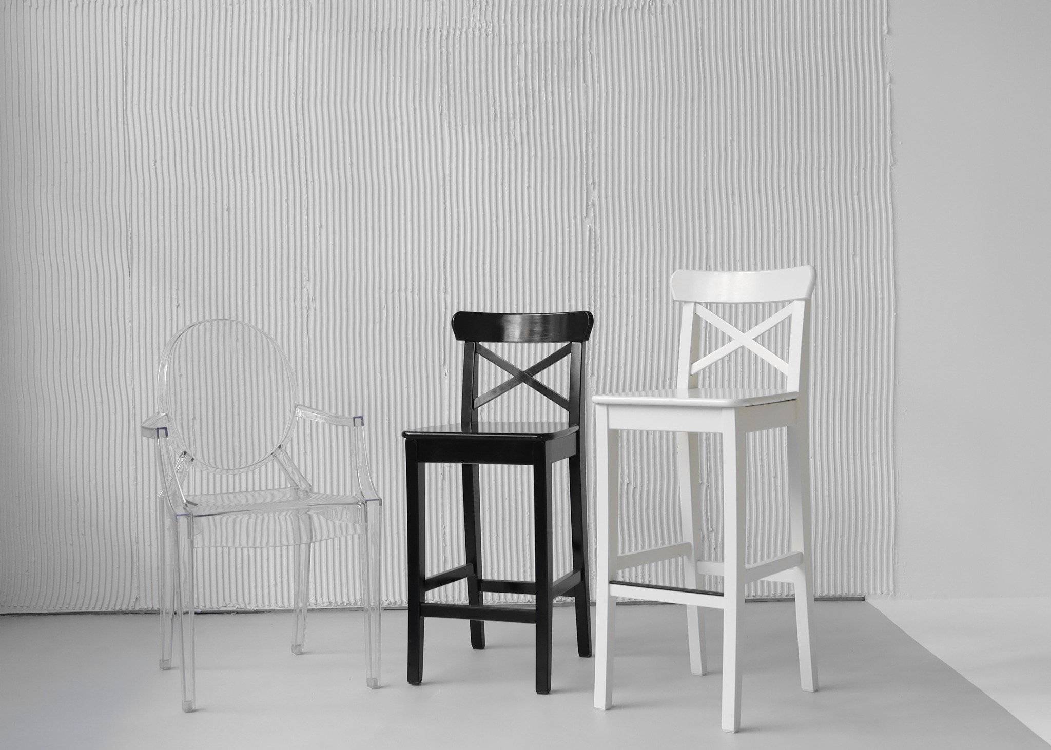 Minimalistyczne ujęcie trzech krzeseł barowych: przezroczystego akrylowego krzesła z podłokietnikami, czarnego drewnianego krzesła z krzyżowym oparciem i białego drewnianego krzesła z krzyżowym...