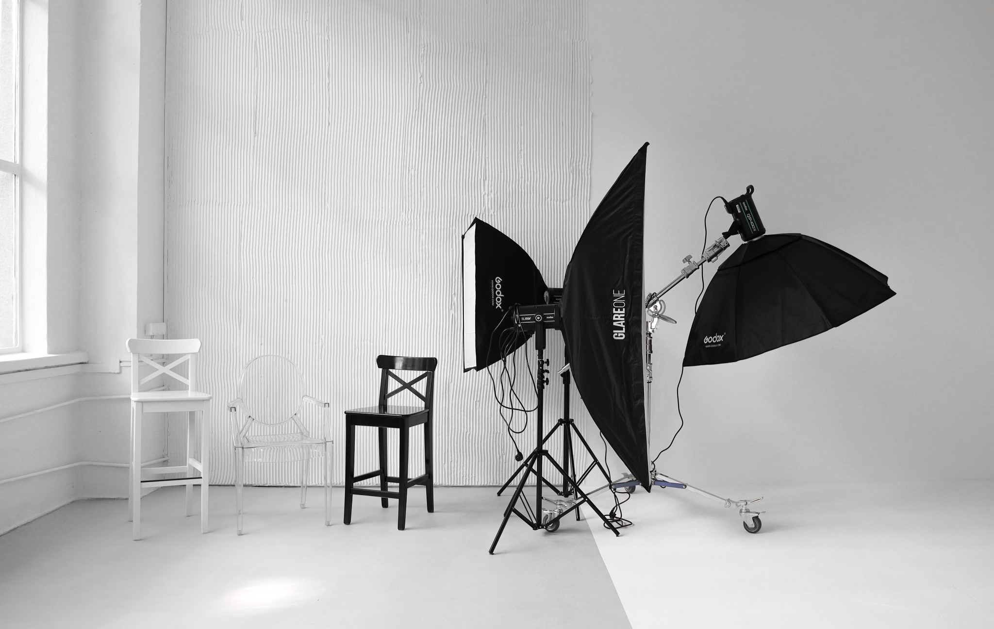 Studio fotograficzne z trzema krzesłami (białym, przezroczystym i czarnym) oraz zestawem oświetleniowym: softboxy Godox i GlareOne, lampy błyskowe na statywach z kółkami, na tle białej ściany...