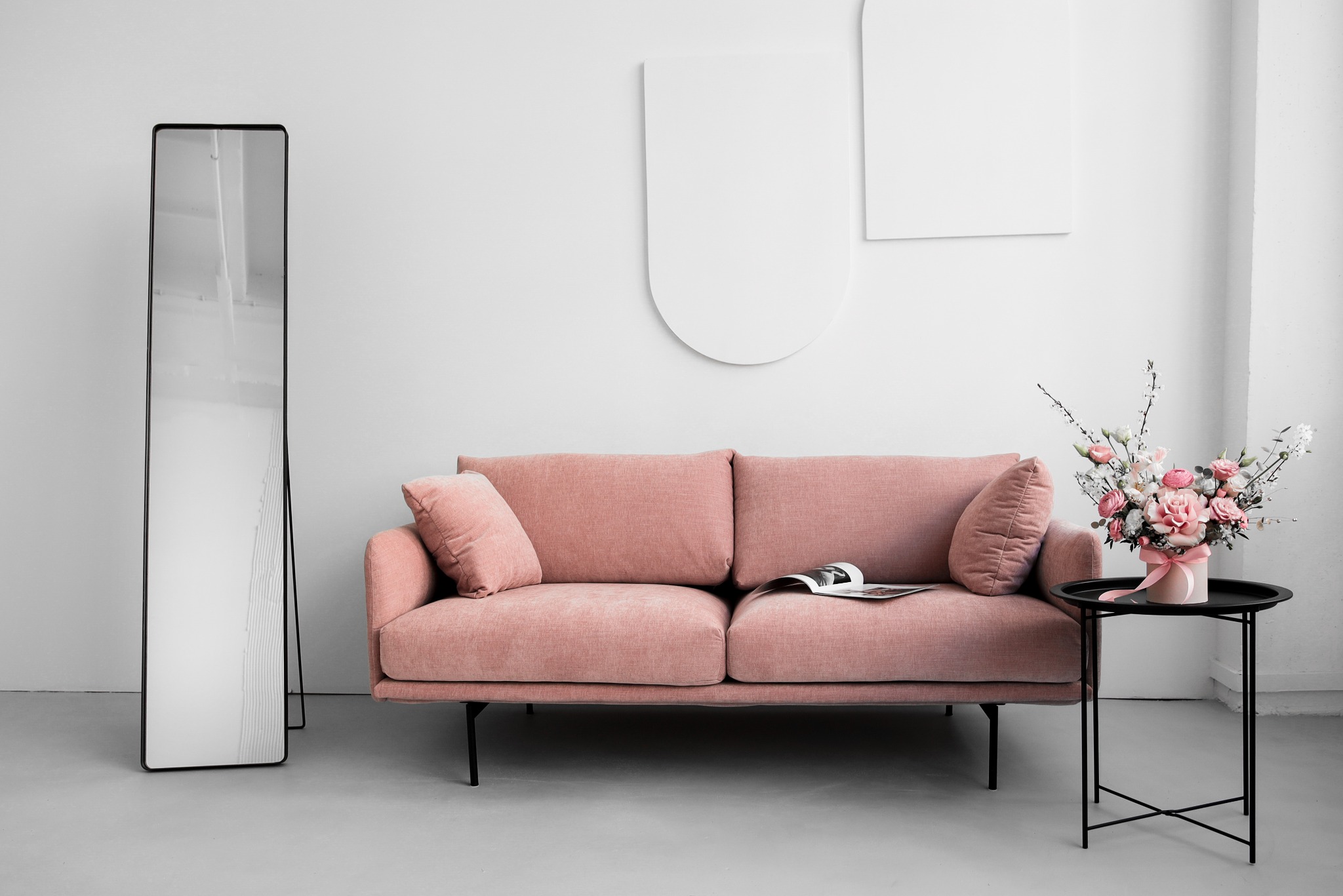Jasnoróżowa kanapa z poduszkami, czarny stolik z bukietem różowych róż, wysokie lustro i minimalistyczne dekoracje ścienne w jasnym, minimalistycznym wnętrzu.