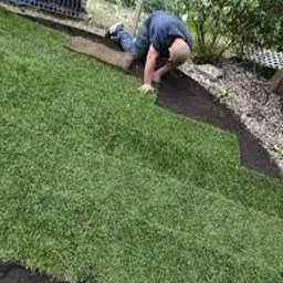 Dom i ogród Usługi Krystian Kłonowski - Mężczyzna na kolanach układa rolkę trawy na przygotowanym podłożu z czarnej tkaniny. Widoczny fragment kolejnej rolki i narzędzia w tle.