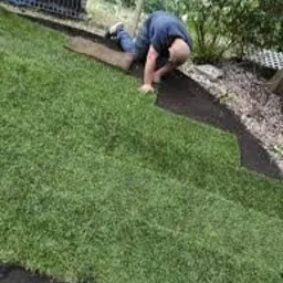 Mężczyzna na kolanach układa rolkę trawy na przygotowanym podłożu z czarnej tkaniny. Widoczny fragment kolejnej rolki i narzędzia w tle.