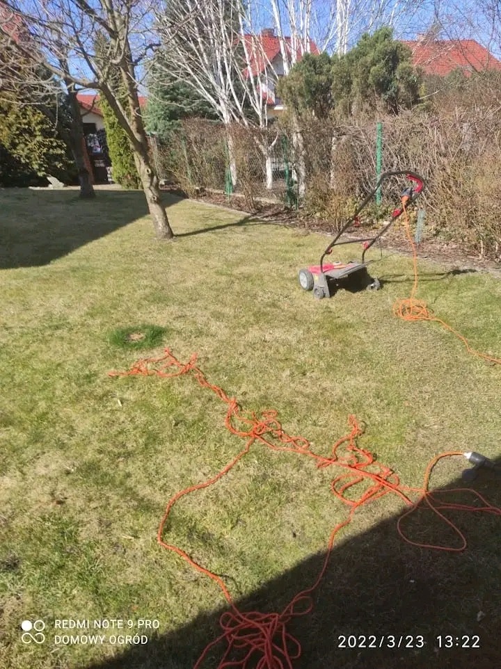 Elektryczna kosiarka do trawy na nierówno skoszonej, zielonej trawie, z pomarańczowym, splątanym kablem zasilającym rozłożonym na trawniku w słoneczny dzień.