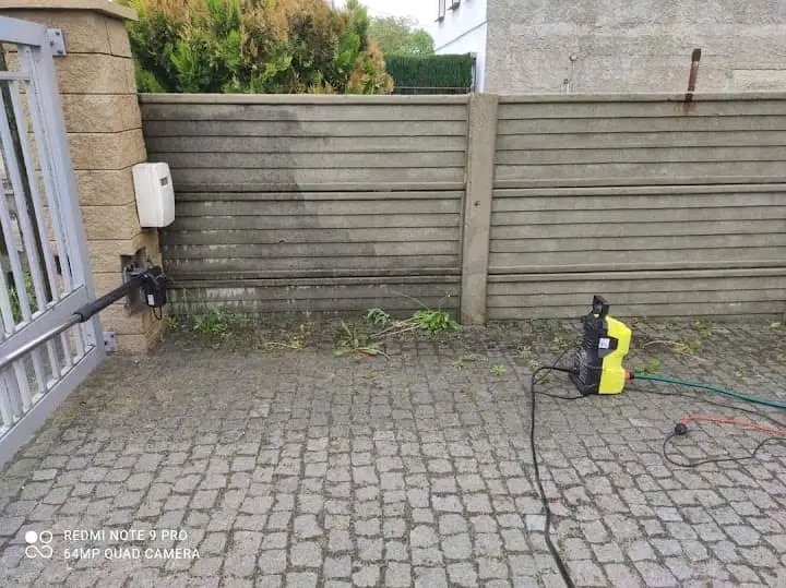 Czyszczenie betonowego ogrodzenia myjką ciśnieniową; widoczna różnica w czystości między umytą a nieumytą częścią, automat do bramy przy słupku.