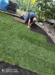 Mężczyzna na kolanach układa rolkę trawy na przygotowanym podłożu z czarnej tkaniny. Widoczny fragment kolejnej rolki i narzędzia w tle.