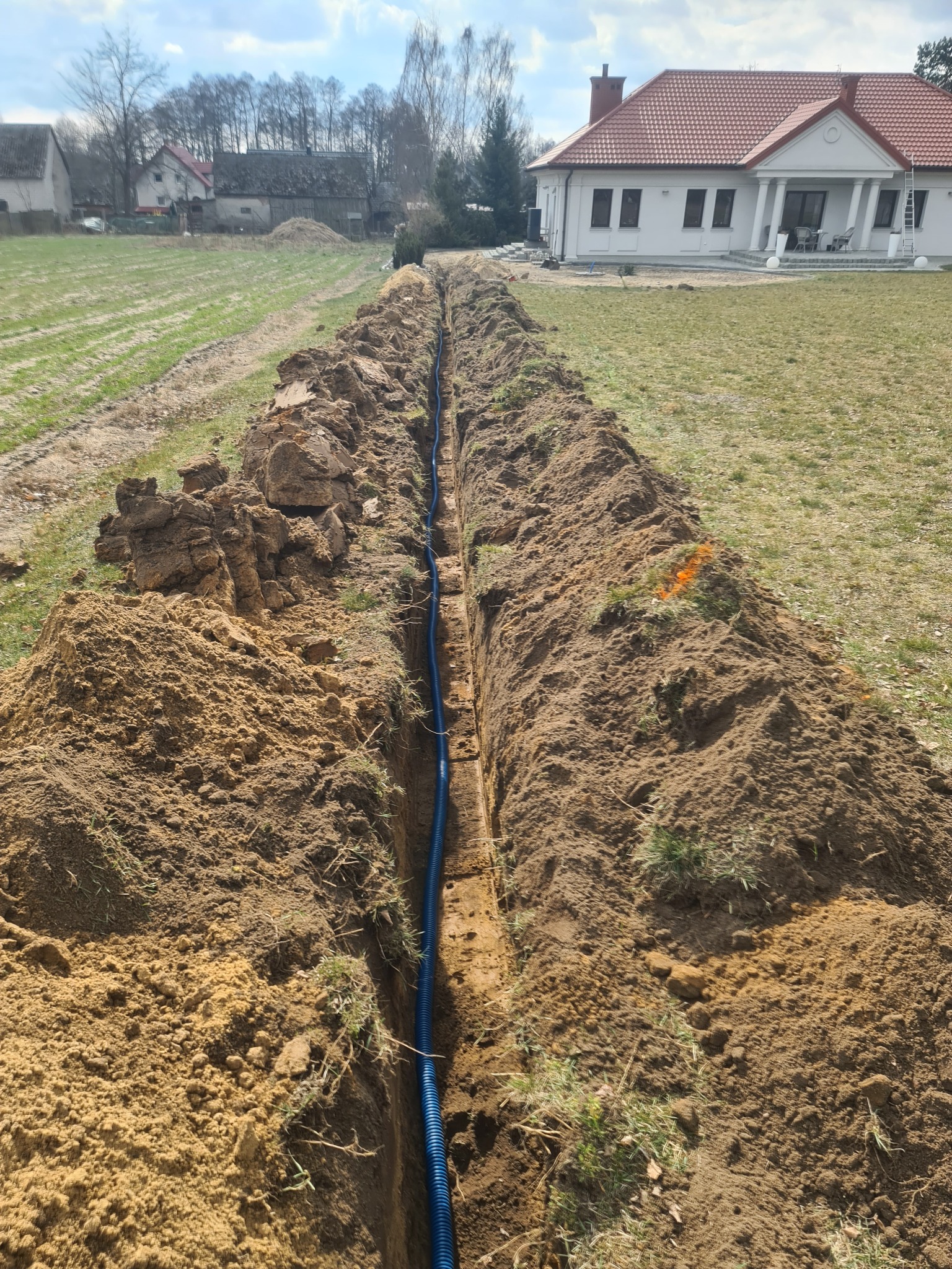 Wykopany rów z niebieską rurą karbowaną na dnie, obok pryzmy ziemi, w tle dom jednorodzinny z czerwoną dachówką i zabudowania gospodarcze.