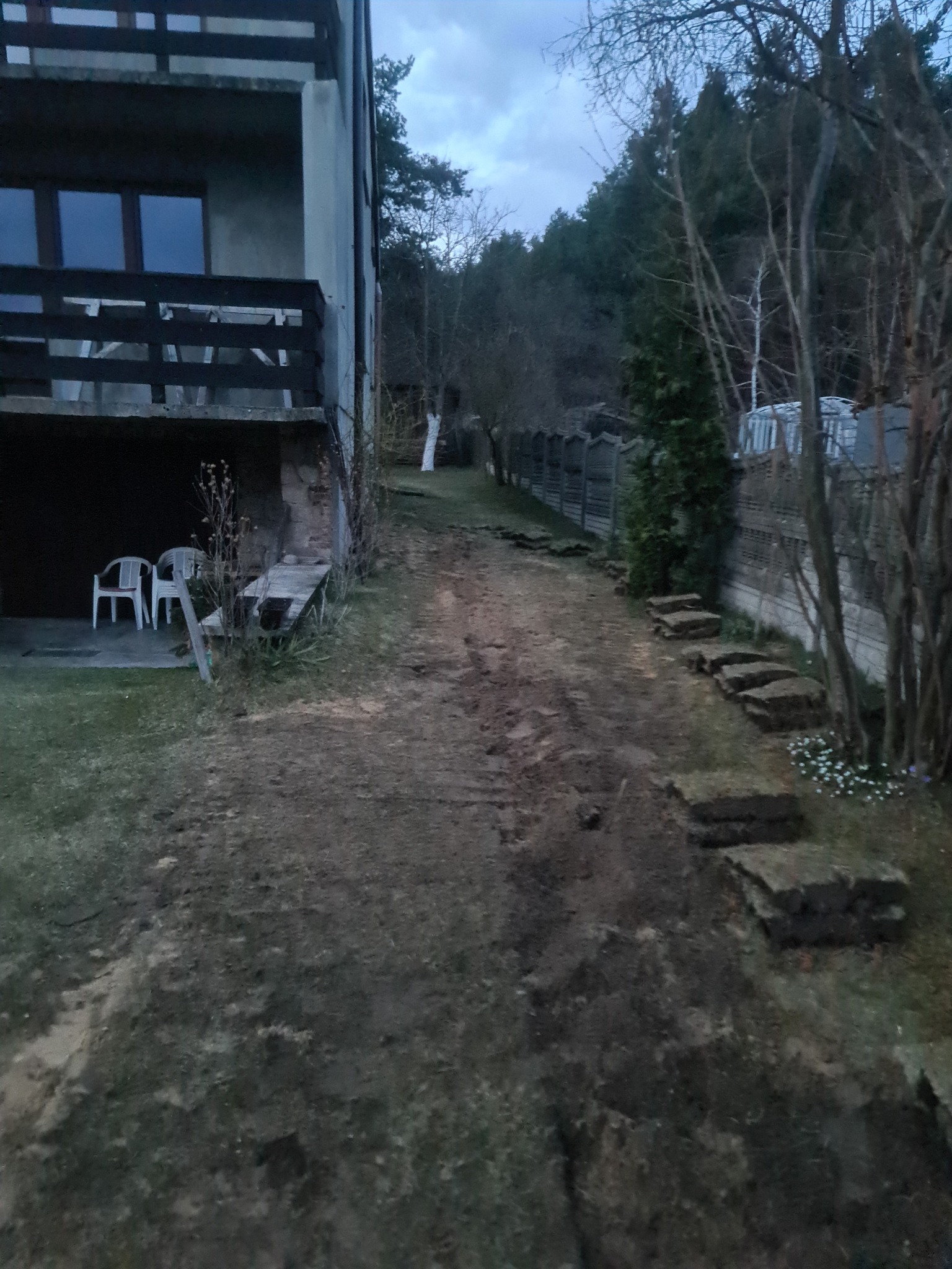 Przygotowany teren pod budowę z widocznymi wykopami pod fundamenty, obok budynku mieszkalnego z balkonem i ogrodem w tle.