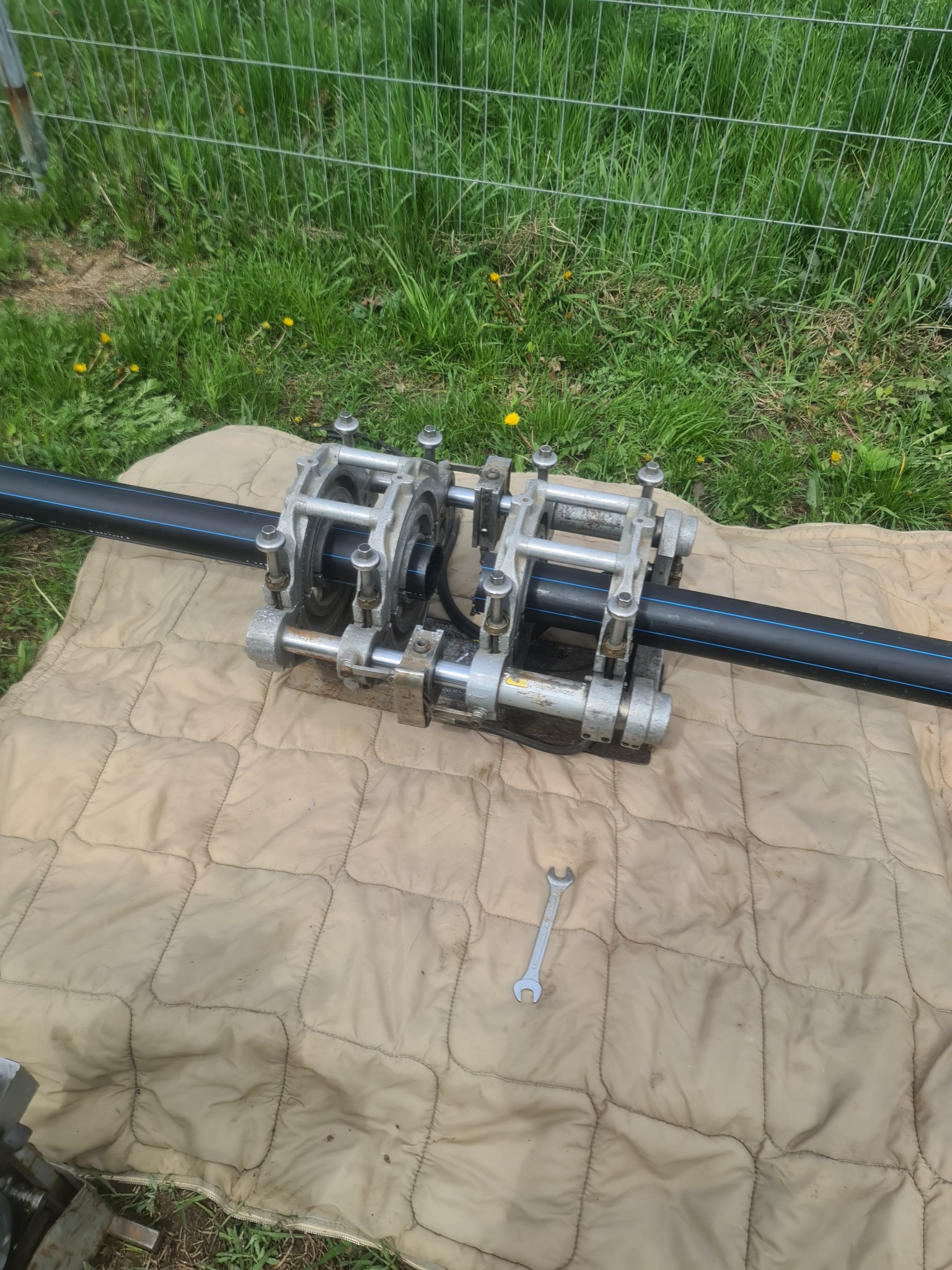 Zgrzewarka do rur PE, z kluczem płaskim obok, leżąca na brudnym kocu w trawie przy ogrodzeniu.