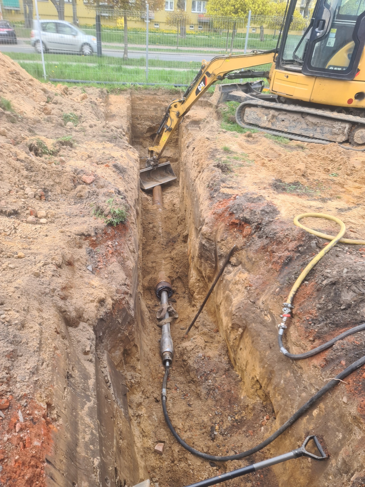 Żółta koparka CAT umieszcza rurę w głębokim, wąskim wykopie w ziemi, z widocznymi narzędziami i przewodami na krawędzi.