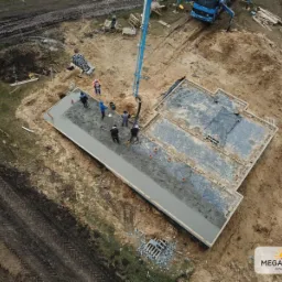 Zalewanie betonem szalunku fundamentowego pod budynek jednorodzinny, widok z drona, ekipa budowlana rozprowadza beton, w tle pompa do betonu i teren budowy.