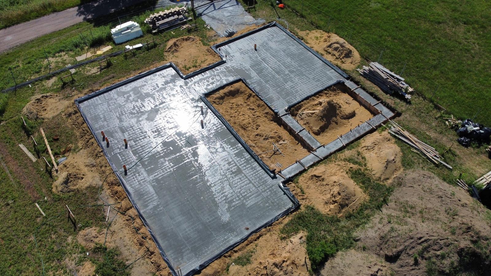Świeżo wylany beton fundamentowy z widocznymi pionowymi rurami instalacyjnymi, częściowo zaszalowany, z perspektywy lotniczej, z widocznymi stertami piasku i resztkami materiałów budowlanych w tle.