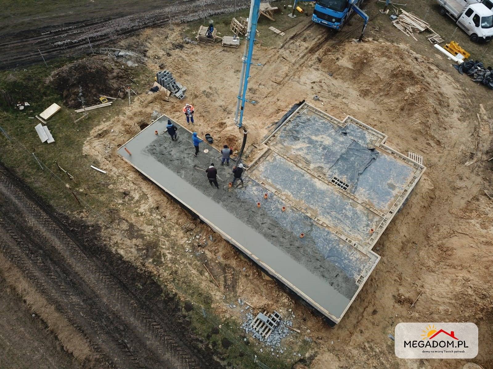 Zalewanie betonem szalunku fundamentowego pod budynek jednorodzinny, widok z drona, ekipa budowlana rozprowadza beton, w tle pompa do betonu i teren budowy.