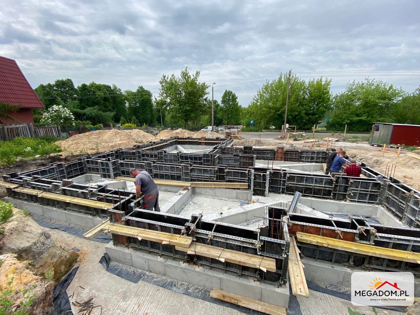 Budowa fundamentów domu jednorodzinnego: pracownicy w szarych ubraniach pracują przy szalunkach zalanych betonem, widoczne zbrojenia i drewniane elementy konstrukcyjne, w tle dom z czerwoną...