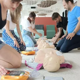 Grupa osób ćwiczy udzielanie pierwszej pomocy na fantomach, używając defibrylatorów AED w jasnym, nowoczesnym pomieszczeniu.