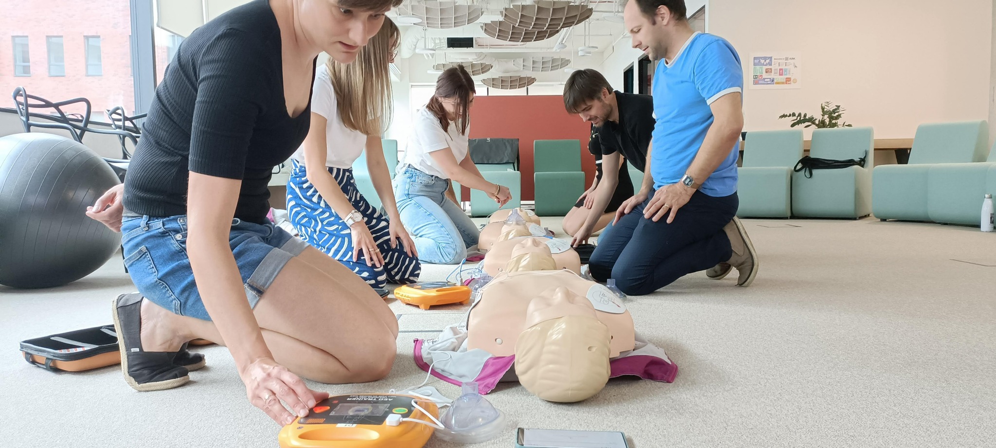 Grupa osób ćwiczy udzielanie pierwszej pomocy na fantomach, używając defibrylatorów AED w jasnym, nowoczesnym pomieszczeniu.