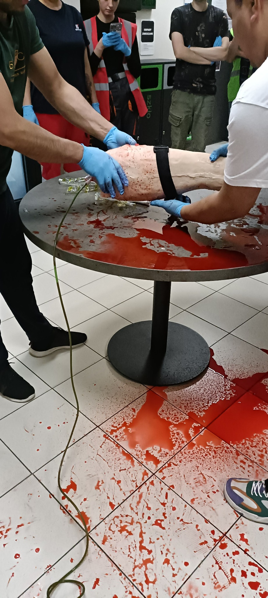 Instruktorzy w niebieskich rękawiczkach opatrują symulowany uraz nogi manekina na metalowym stole; czerwona ciecz rozlana na stole i podłodze z płytek.