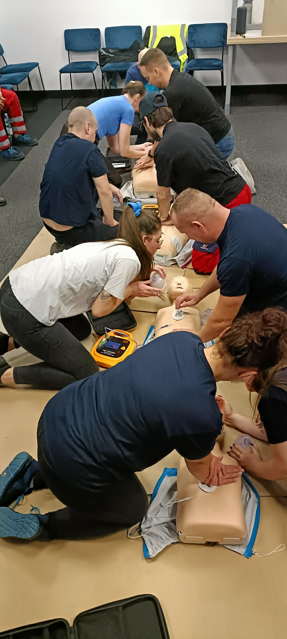 Grupa osób ćwiczy resuscytację krążeniowo-oddechową na manekinach, używając masek resuscytacyjnych i defibrylatora AED Trainer. Widoczny instruktor w czerwonym stroju ratowniczym.