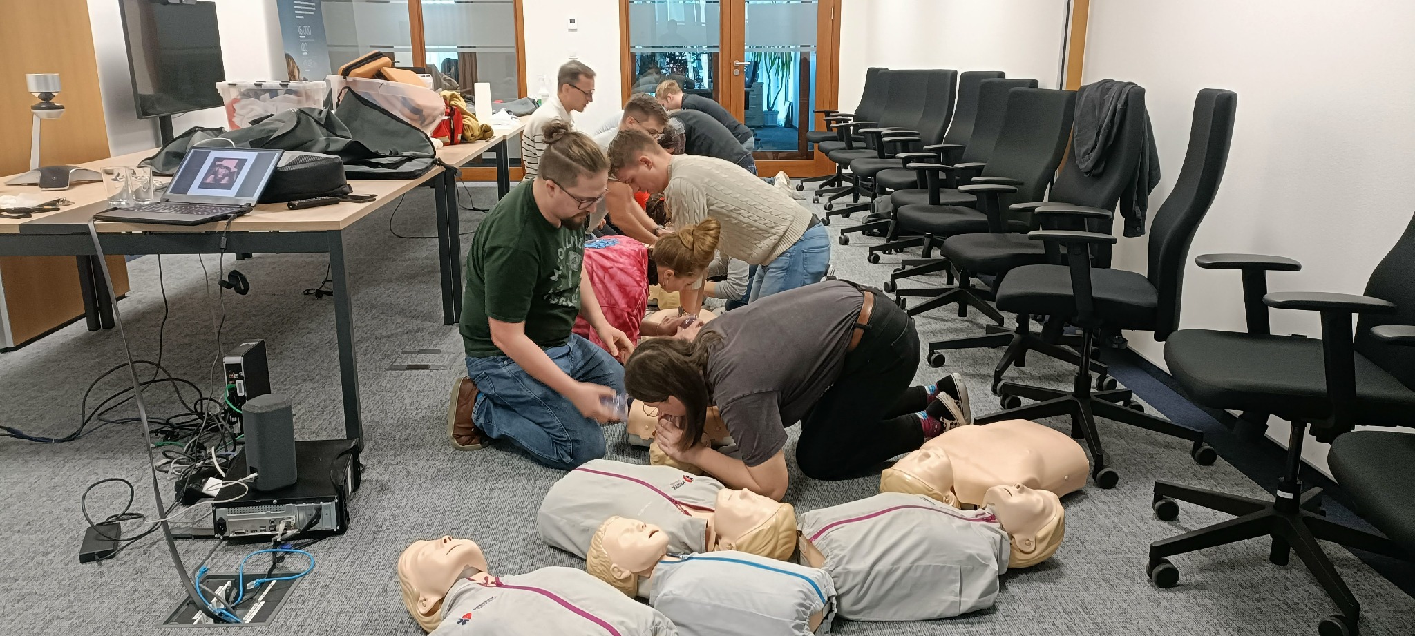 Grupa osób ćwiczy resuscytację krążeniowo-oddechową na manekinach w sali konferencyjnej z rzędem czarnych krzeseł w tle.